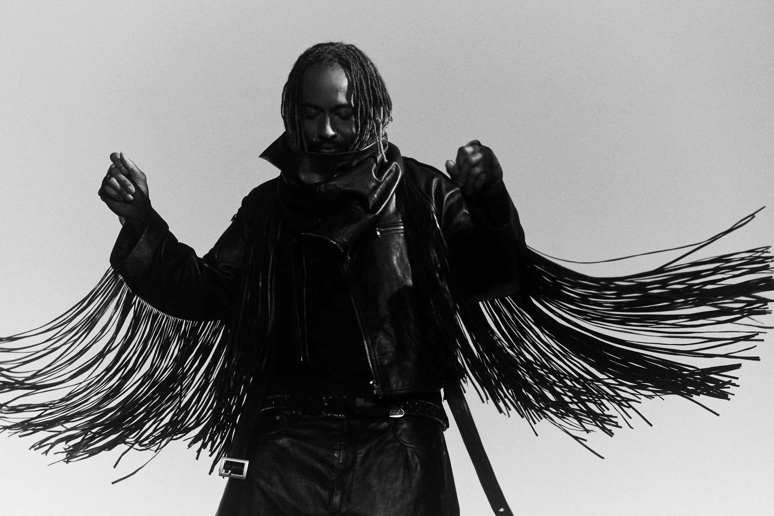 Black and white portrait of Thundercat wearing a fringed leather jacket with arms raised, shot from below against an open sky